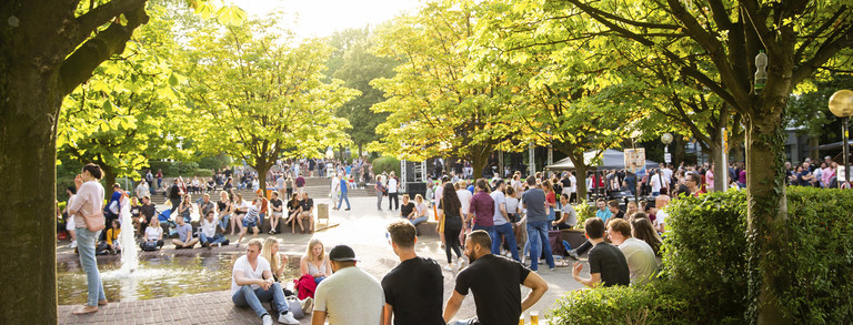 Viele Personen halten sich rund um einen Brunnen auf. Dieser ist umgeben von Bäumen.