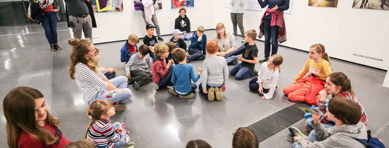 Kinder sitzen auf dem Boden, eine Frau spricht zu ihnen.