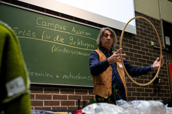 A man stands in front of a blackboard holding a wooden hoop in his hands.