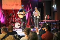 A woman sings and a man plays guitar and both are on a small stage in front of an audience. 