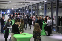 Photo: Several people stand and converse at green standing tables in a brightly lit room with large windows.