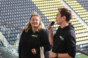 Ein Mann, der ein Mikrofon in der Hand hält, und eine Frau stehen vor einer Tribüne in einem Fußballstadion.