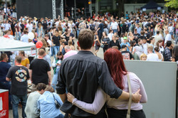 Photo: A couple stand arm in arm at TU Dortmund University's summer festival and watch the hustle and bustle on campus. The couple have their backs to the camera. The stage and people at the summer festival can be seen in the background.