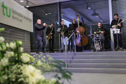 Photo: A music group performs with trumpet, saxophone, double bass, drums, and guitar on a staircase in a building of TU Dortmund University.