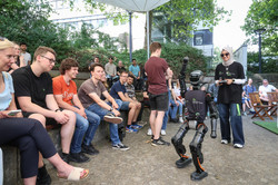 Photo: A woman with sunglasses and a headscarf controls a robot. A curious audience looks on.