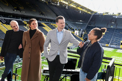 Four people are standing in the BVB stadium stands. One of the people is speaking into a microphone, while the others are looking at her.