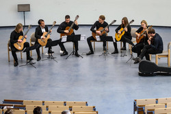 Sieben Gitarrenspieler*innen sitzen in einem Hörsaal mit Notenständern und spielen Musik.