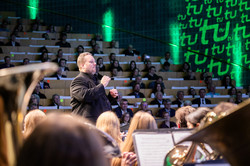 Im Fokus des Bildes ist ein Dirigent zu sehen, welcher gerade ein Orchester leitet, welches in Teilen im Vordergrund zu sehen ist. Im Hintergrund ist das Audimax der TU Dortmund erkennbar sowie das in den Bänken sitzende Publikum. Die Wände sind grün und teilweise mit TU-Logo beleuchtet.