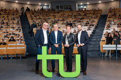 Foto: Vier Personen posieren im Hörsaal der Technischen Universität Dortmund hinter einem großen grünen TU-Logo, während zahlreiche Zuhörende in den Reihen sitzen.