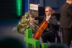 Das Bild zeigt Violoncello-Solist Hauke Hack mit Sonnenbrille, welcher beim Sinfonischen Blasorchester der TU Dortmund beiwohnte. Im Vordergrund ist das TU-Logo erkennbar. Im Hintergrund befindet sich ein Pult mit TU-Schild und einem Blumengesteck davor.