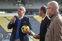 Three men, one of whom is holding a ball and a microphone, stand side by side in front of a stand in a soccer stadium.