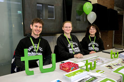 The picture shows a row of white tables in the foreground with lots of information material about TU Dortmund University. Behind the row of tables sit three people in their black university hoodies.