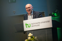 Foto: Eine Person steht am Pult der Technischen Universität Dortmund mit Laptop und Mikrofon, davor ist ein Blumenarrangement zu sehen.