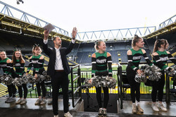 Eine Gruppe von Cheerleaderinnen auf einer Stadiontribüne.Sie halten silber-schwarze Ponpons und formen eine La-Ola Welle. In der Mitte steht ein Moderator.