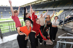 Eine Gruppe sportlicher gekleideter Menschen, die für ein Foto posieren. Sie stehen auf eine Stadiontribüne. Im Hintergrund sieht man das BVB-Stadion.