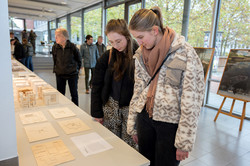 The photo shows a long table with wooden models. Visitors are looking at them. A bright window front and pictures on easels can be seen in the background.