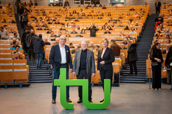 Prof. Manfred Bayer, Prof. Klaus von Klitzing and Evi Hoch (from left to right) in front of the Audimax. In front of them is a green TU logo.
