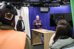 Ein Mann hält einen Vortrag in einem Fernsehstudio. Im Vordergrund sieht man Menschen im Publikum.