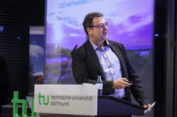 Photo: A person stands at the podium of TU Dortmund University and points to a presentation in the background.