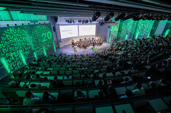 Das Bild zeigt das Audimax der TU Dortmund bei der Akademischen Jahresfeier 2024. Das Foto wurde von oben aufgenommen und zeigt das Publikum von hinten. Im Zentrum, weiter oben im Bild ist das Orchester der TU Dortmund zu sehen, darüber die Präsentation. Die seitlichen Wände des Audimax sind grün angestrahlt und werden zusätzlich auf beiden Seiten mit zwei Kreisen, in denen das TU-Logo sowie die Zahl 24 angezeigt wird, angestrahlt.