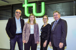 Group photo: Four people stand in a room in front of a large, brightly lit green TU logo.