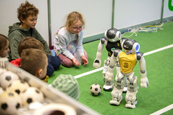 The picture shows two robots in the corner of a soccer pitch, looking at the ball. Behind the line, children are sitting on the ground watching the game.