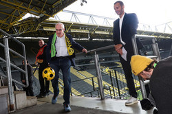 Prof. Manfred Bayer schießt einen schwarz-gelben Ball. Dabei steht er auf einer Stadiontribüne. Neben ihm stehen mehrere Männer.
