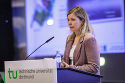Foto: Eine Person spricht am Rednerpult der Technischen Universität Dortmund vor einer Leinwand mit einer Präsentation.