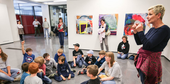 KinderUni-U Kinder sitzen in einem Kreis auf dem Boden. An der Wand hinter ihnen hängen Bilder. Eine Frau dolmetscht für Gehörlose.