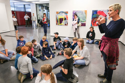 KinderUni-U Kinder sitzen in einem Kreis auf dem Boden. An der Wand hinter ihnen hängen Bilder. Eine Frau dolmetscht für Gehörlose.