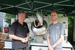 Photo: Two men stand at a stand at TU Dortmund University's summer festival and smile into the camera. Behind them is a large apparatus for an experimental setup.