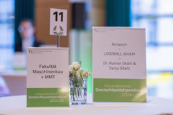 Aufsteller auf einem Stehtisch weisen darauf hin, dass hier die Fakultät Maschinenbau sowie die Fördernden von Amazon, der Logiball GmbH sowie die privaten Fördernden Dr. Rainer Stahl und Tanja Stahl mit Ihren Stipendiat*innen zusammenkommen können