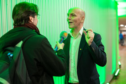 A man with a backpack stands in front of another man and holds a microphone out to him.
