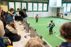 Auf einer grünen Kunstrasenfläche spielen zwei kleine, schwarze Roboter gegeneinander Fußball, betreut von drei Studenten mit schwarzen T-Shirts, die im Hintergrund stehen. Links sitzt und steht das Publikum an Tischen mit Computerbildschirmen.