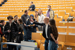 Several people are standing in a lecture hall. In the foreground, a man shoots a selfie with a young woman using a cell phone.