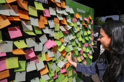 Photo: A woman hangs a Post-It on a green wall with many colorful Post-Its stuck to it.