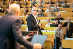 Foto: Eine Person steht mit Mikrofon vor einem Publikum in einem Hörsaal, während eine andere Person im Vordergrund am Pult mit einem Laptop steht.