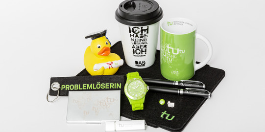 Verschiedene Merchandise-Artikel der TU Dortmund, unter anderem eine Tasse, eine Uhr und ein Schlüsselanhänger, liegen auf einem Tisch