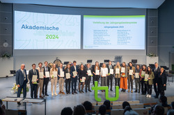 Das Bild zeigt ein Gruppenfoto der Personen mit den besten Abschlussarbeiten im Jahr 2024. In der Mitte vor den Personen steht ein großes, grünes TU-Logo. Im Vordergrund sind dunkel die Umrisse des Publikums erkennbar. Im Hintergrund sind ebenso zwei Präsentationsbilder zu sehen.