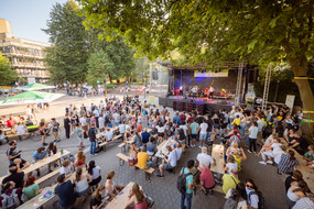 In front of a music stage, many people are gathered standing and sitting on beer tent benches.