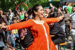 Photo: A woman with dark hair and an orange dress dances with her arms outstretched at the summer festival. In the background, people sit and stand in the audience.