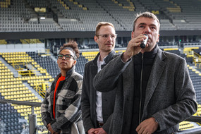 Two men, one of whom is holding a microphone, and a woman are standing in front of a stand in a soccer stadium.