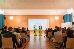 Blick aus den hinteren Publikumsreihen durch den Mittelgang nach vorne zum Bühnenbereich, wo Moderatorin Britt Lorenzen gerade mit Rektor Prof. Dr. Manfred Bayer im Gespräch ist