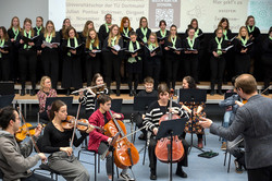 The photo shows the university choir in black clothing with a light green scarf. The university orchestra can be seen in the foreground.