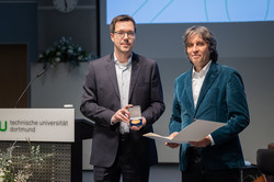 Das Bild zeigt Jan Nagel und Preisträger Schwentick, die in die Kamera lächeln. Jan Nagel hält eine Schachtel mit Medaille in der Hand und Preisträger Schwentick eine Urkunde. Links von den Personen sieht man das Pult mit TU-Schild und einen Teil des Blumengestecks, welches sich vor dem Pult befindet..