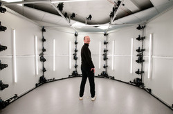 The picture shows a male person dressed in black in the middle of a white, circular room, turning his upper body backwards to smile at the camera. Cameras hang on the wall and ceiling, evenly spaced one above the other.