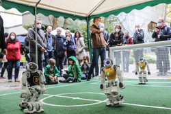  Several people are standing around a small green soccer field where three small white robots are playing.