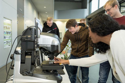 In the center of the picture is a device standing on a table. Four other people are looking at the device.