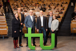 Four men and two women are standing in front of the two green letters t and u. In the background you can see a lecture hall where several people are sitting.
