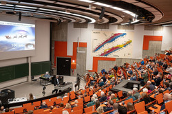 In einem gut besuchten Hörsaal mit orangenen Sitzen hält Prof. Metin Tolan einen Vortrag. Auf der Leinwand ist eine Präsentation zu sehen.